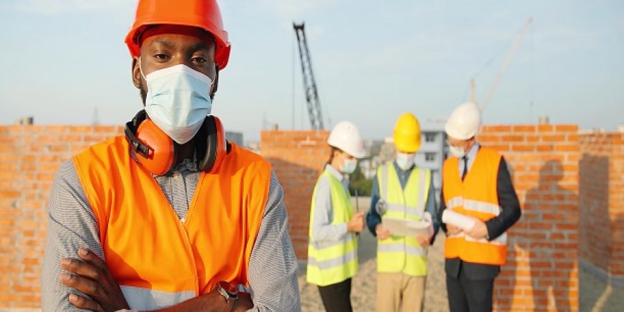 Portrait of African American handsome young man constructor in casque and medical mask standing outdoor at construction and looking at camera. Male builder at building top in helmet. Coronavirus. Portrait of African American handsome young man constructor in casque and medical mask standing outdoor at construction and looking at camera. Male builder at building top in helmet.