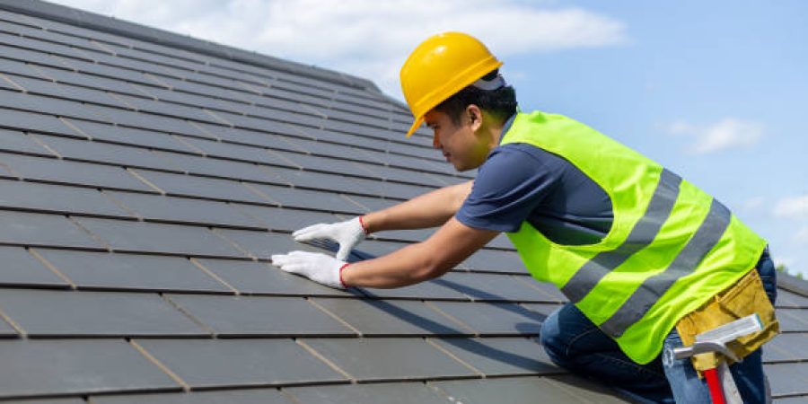 Level 2 NVQ Diploma in Roofing Occupations Roof repair, worker with white gloves replacing gray tiles or shingles on house with blue sky as background and copy space, Roofing - construction worker standing on a roof covering it with tiles.